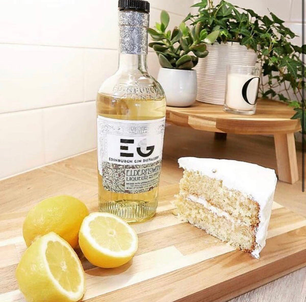 Picture shows, lemon and two sliced lemons, slice of lemon cake, bottle of Edinburgh lemon and elderflower gin, plants in background, white tiles on wall